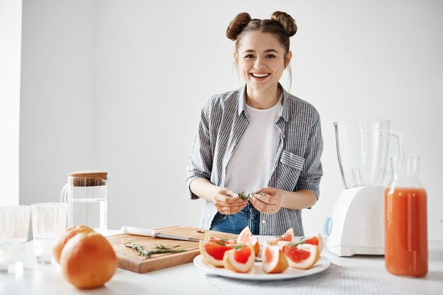 Jolie fille souriante plaque de décoration pour le petit déjeuner avec des tranches de pamplemousse et de romarin sur mur blanc. Smoothie détox rafraîchissant.