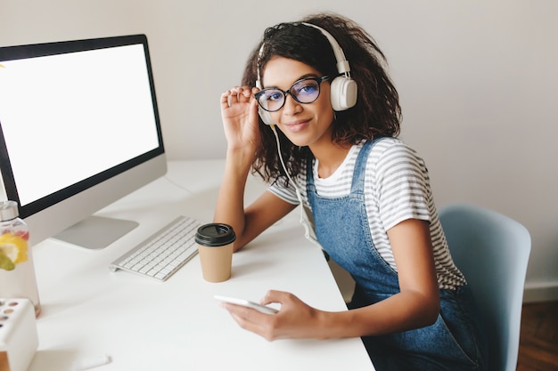 Photo gratuite jolie fille avec un sourire fatigué posant sur le lieu de travail près de l'ordinateur avec écran blanc