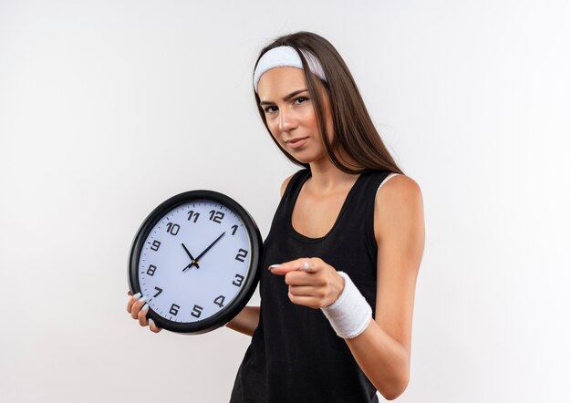 Jolie fille sportive confiante portant un bandeau et un bracelet tenant une horloge et pointant isolé sur un mur blanc