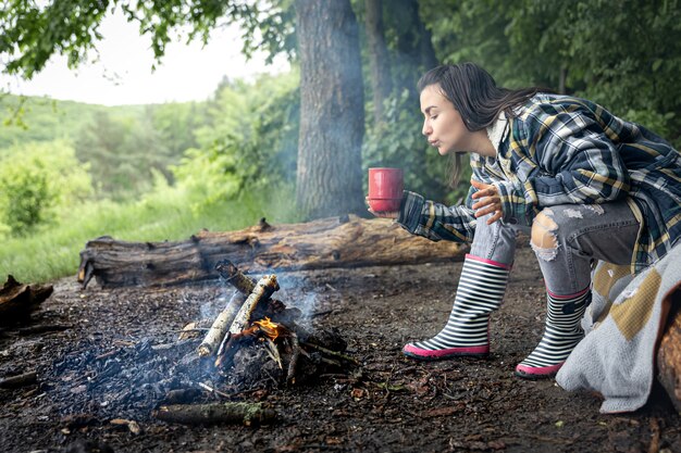 Une jolie fille avec une tasse à la main se réchauffe près d'un feu dans la forêt.