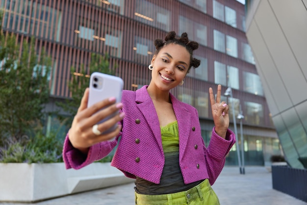 Une jolie fille en tenue à la mode crée un contenu d'influence montre un signe de paix à la caméra frontale du smartphone pose pour des sourires de selfie pose avec plaisir dans un lieu urbain passe des vacances dans une grande ville.