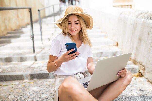 Jolie fille travaillant avec ordinateur portable et téléphone assis sur les escaliers de la rue de la ville.