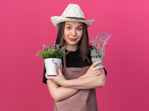 Une jolie jardinière caucasienne heureuse portant un chapeau de jardinage se tient avec les bras croisés tenant des pots de fleurs isolés sur un mur rose avec un espace pour copie