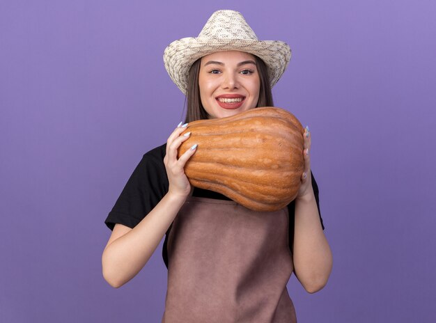 Une jolie jardinière caucasienne souriante portant un chapeau de jardinage tient une citrouille