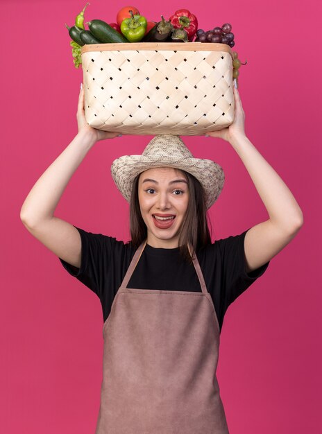 Jolie jardinière caucasienne surprise portant un chapeau de jardinage tenant un panier de légumes au-dessus de la tête isolée sur un mur rose avec espace pour copie