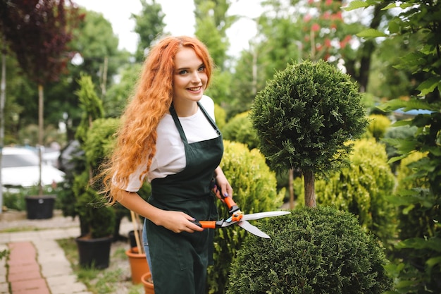 Jolie jardinière souriante aux cheveux bouclés roux debout dans un tablier et tenant de gros ciseaux de jardin tout en regardant joyeusement à huis clos à l'extérieur
