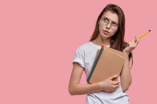 Jolie jeune adolescente a une expression réfléchie, un regard attrayant, tient le bloc-notes en spirale avec un crayon, pense à l'écriture d'un essai