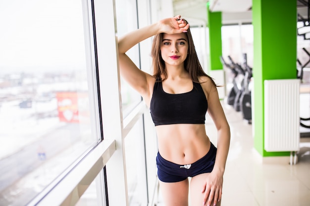 Jolie jeune beauté aux grands yeux verts corps en forme solide longs cheveux bruns pose dans la salle de gym devant les fenêtres