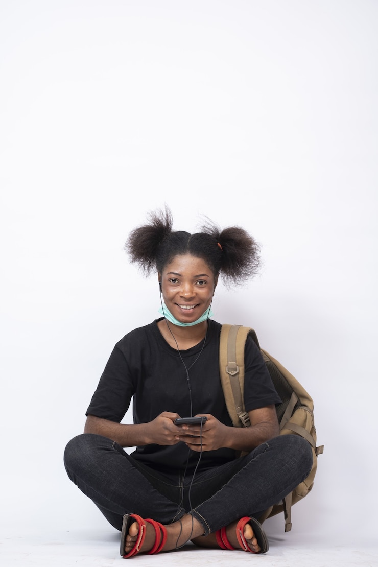 Photo gratuit jolie jeune femme africaine portant un sac à dos, les jambes assises croisées à l'aide de son téléphone portable