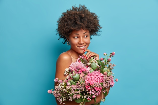 Jolie jeune femme afro-américaine réfléchie se tient nue à l'intérieur tient un grand bouquet de fleurs regarde pensivement et se mord les lèvres