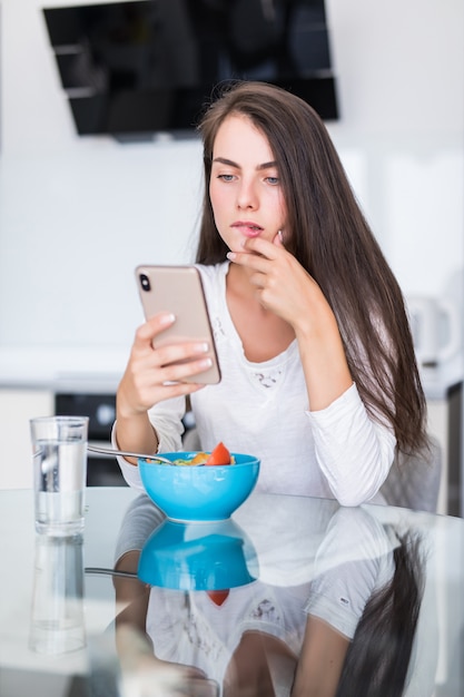 Jolie jeune femme à l'aide de son téléphone portable tout en mangeant de la salade dans la cuisine à la maison.