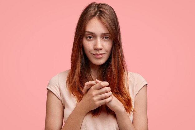 Jolie jeune femme avec une apparence agréable, garde les mains jointes et regarde mystérieusement la caméra, isolée sur fond rose.