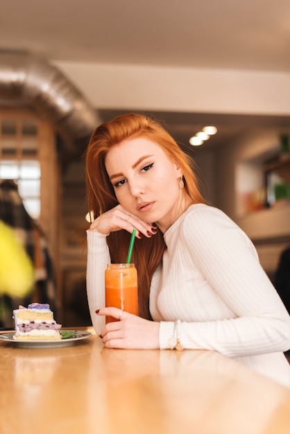 Une jolie jeune femme assise au café avec une délicieuse tranche de gâteau et un verre à smoothie sur une surface en bois