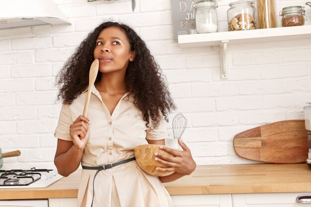 Jolie jeune femme au foyer afro-américaine en robe beige debout dans la cuisine avec des ustensiles et une cuillère en bois ayant une expression faciale pensive, pensant quoi cuisiner pour le dîner. Cuisine et nourriture