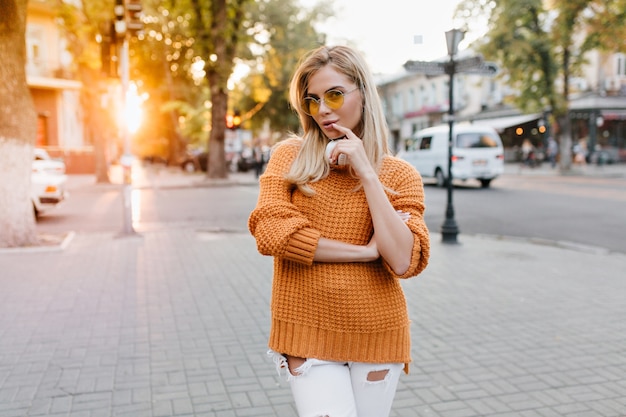 Jolie jeune femme aux cheveux blonds brillants posant sur la place de la ville avec l'expression du visage heureux