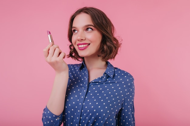 Jolie jeune femme aux cheveux bouclés isolé sur mur rose avec rouge à lèvres à la main. fille adorable souriante porte chemisier bleu.