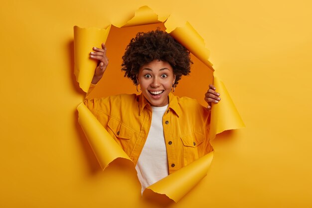 Jolie jeune femme aux cheveux bouclés s'amuse, pose dans un trou de papier,