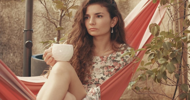 Jolie jeune femme aux cheveux roux en robe à fleurs lit un livre dans un hamac dans un petit jardin patio urbain