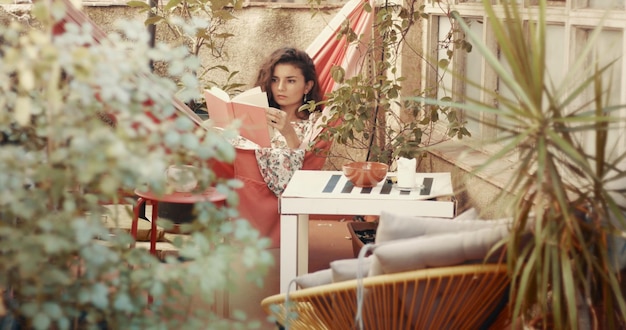 Jolie jeune femme aux cheveux roux en robe à fleurs lit un livre dans un hamac dans un petit jardin patio urbain