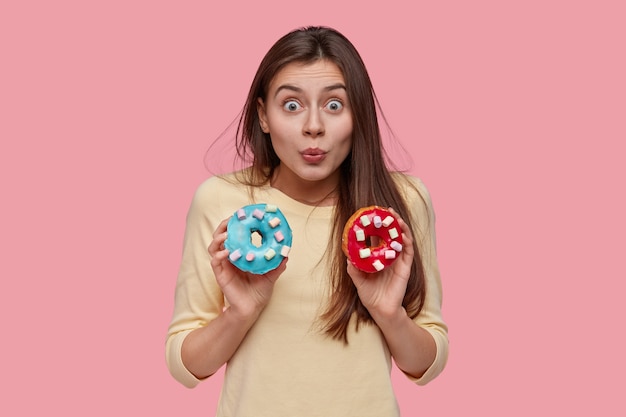 Jolie jeune femme aux longs cheveux noirs, tient des beignets tasy, suggère de prendre une collation avec elle, pose sur un espace rose