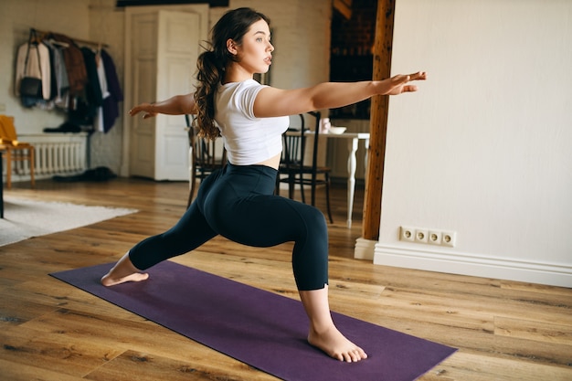 Jolie jeune femme aux pieds nus pratiquant le yoga à la maison, debout sur un tapis, faisant la posture du guerrier II ou Virabhadrasana, renforçant les jambes, ouvrant les hanches et développant la concentration et l'équilibre