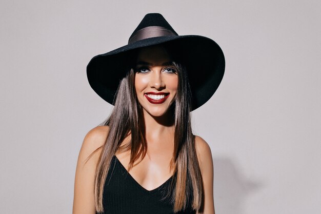 jolie jeune femme aux yeux charbonneux et aux lèvres noires portant un chapeau et une robe noire posant devant le mur. Halloween, mascarade, fête, célébration