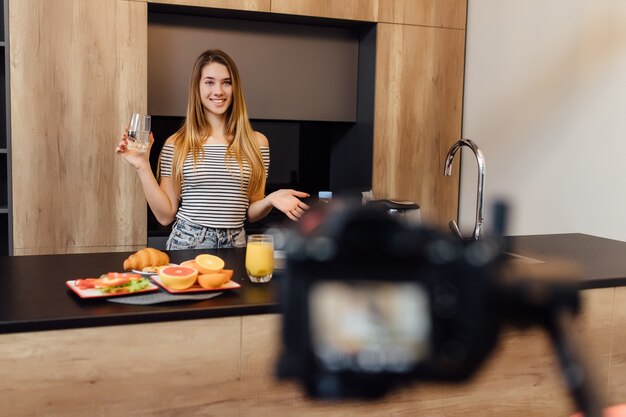 Jolie jeune femme blonde blogueuse de l'eau potable dans la cuisine avec des aliments sains sur la table