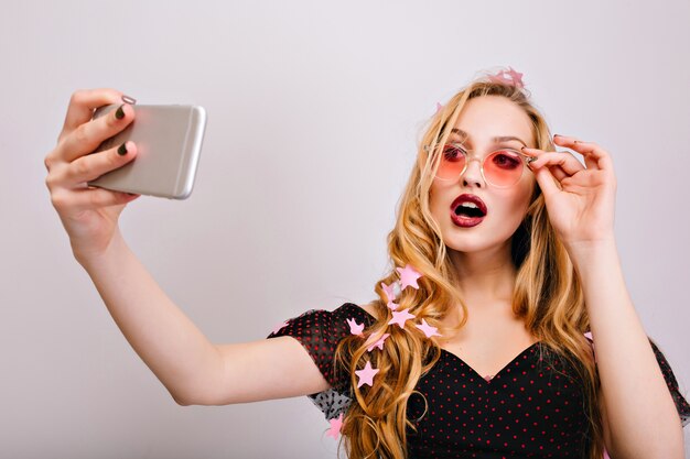 Jolie jeune femme blonde prenant selfie à la fête, faisant un look sexy avec la bouche ouverte. Porter des lunettes roses élégantes, une robe noire, a de beaux cheveux longs bouclés.