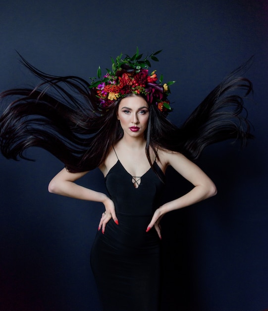 Jolie jeune femme brune aux cheveux longs avec un maquillage lumineux vêtue d'une grande couronne florale colorée sur le fond bleu foncé vêtue d'une robe noire