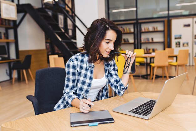 Jolie jeune femme brune étudie avec un ordinateur portable dans la bibliothèque. Étudiant intelligent, vie universitaire, surfant sur internet, occupé, travaillant, exprimant des émotions positives, souriant, jouant avec des lunettes noires.