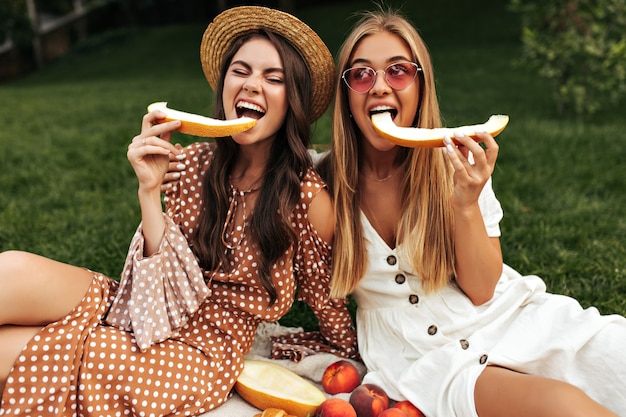 Jolie jeune femme brune et jolie fille blonde bronzée dans des robes d'été élégantes mangent du melon et pique-niquent à l'extérieur