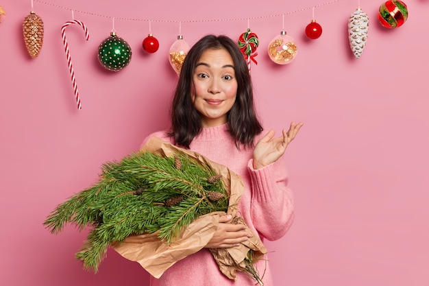 Jolie jeune femme brune a un passe-temps créatif soulève la paume avec hésitation ne sait pas quoi faire des branches d'épinette verte se prépare pour les vacances d'hiver pose sur le décor de Noël à la maison