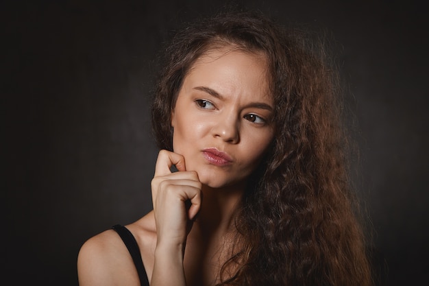 Jolie jeune femme brune soignée et bien entretenue avec de longs cheveux volumineux fronçant les sourcils et regardant de côté, se grattant le visage, réfléchissant à un problème, essayant de trouver une solution.