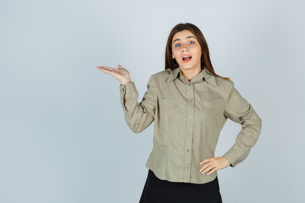 Jolie jeune femme en chemise, jupe faisant semblant de montrer quelque chose en écartant la paume et en se demandant, vue de face.