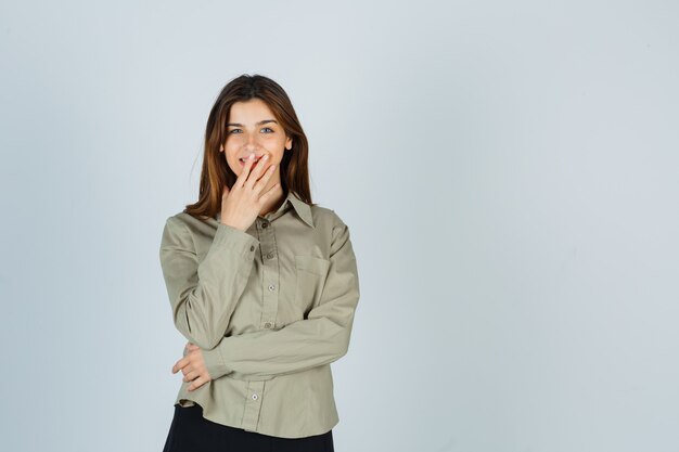 Jolie jeune femme en chemise, jupe tenant la main sur la bouche et l'air joyeux, vue de face.