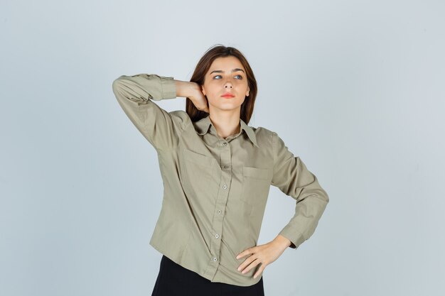 Jolie jeune femme en chemise, jupe tenant la main sur le cou et regardant pensive, vue de face.