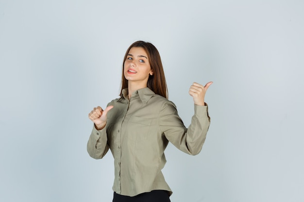 Jolie jeune femme en chemise pointant vers l'arrière avec les pouces et l'air confiant, vue de face.