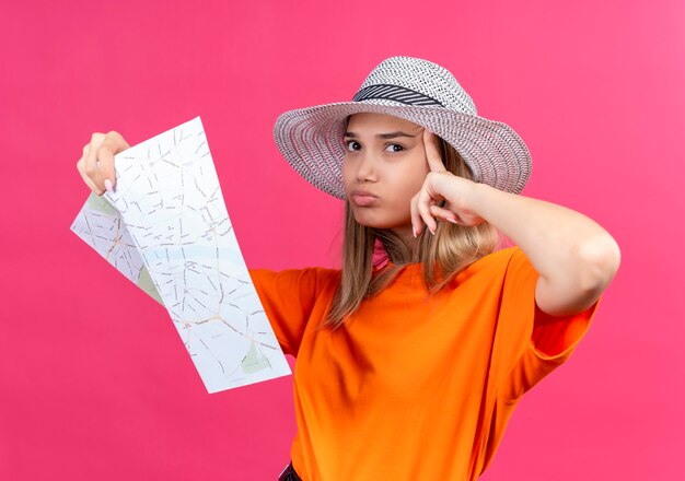 Une jolie jeune femme confuse dans un t-shirt orange portant un chapeau tenant la carte tout en pointant vers la tête et à côté sur un mur rose