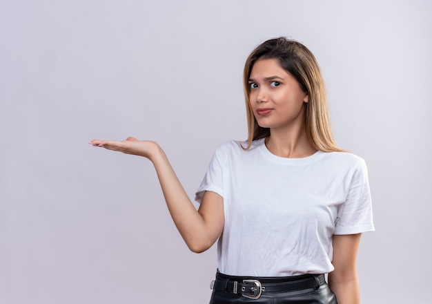 Une jolie jeune femme confuse en t-shirt blanc pointant son bras vers la gauche sur un mur blanc