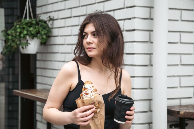 Jolie jeune femme avec un croissant et un café contre un mur de briques blanches