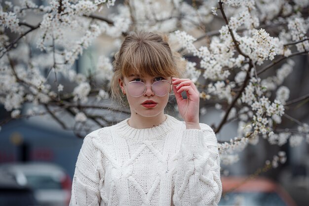 Jolie jeune femme dans un pull blanc dans un jardin de fleurs de cerisier, printemps. photo de mode en plein air