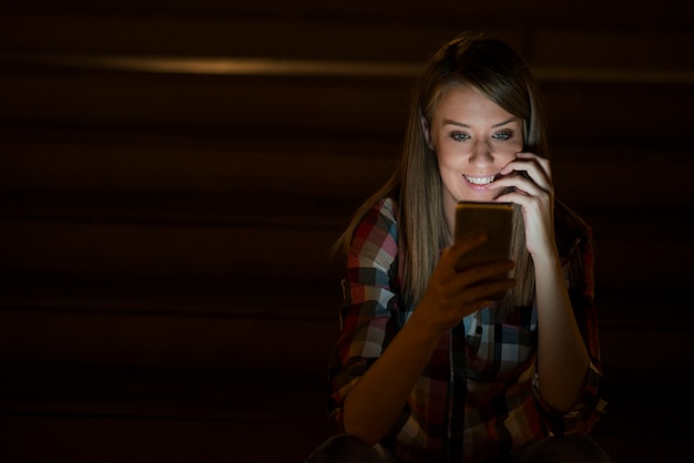 Jolie jeune femme debout avec téléphone mobile dans la rue de nuit à l&#39;extérieur, femme étudiante caucasienne en train de lire des messages texte sur son téléphone portable avec réfléchi sur son écran de visage, téléphone écran vierge