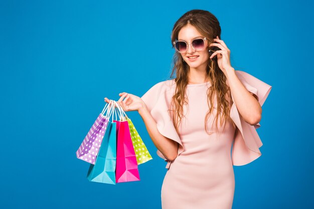 Jolie jeune femme élégante en robe de luxe rose à l'aide d'un téléphone portable et tenant des sacs à provisions