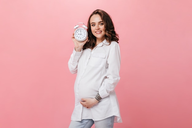 Jolie Jeune Femme Enceinte En Chemise Blanche Sourit Largement Et Pose Sur Isolé. Jeune Fille Brune Détient Un Réveil Sur Fond Rose.