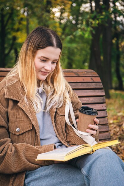Une jolie jeune femme est assise sur un banc dans un parc en automne et lit un livre