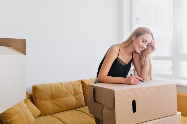 Jolie jeune femme étiquetage boîte en carton, tenant le marqueur à la main, va emballer des choses, déménager dans un nouvel appartement, appartement, maison. Fille heureuse dans la chambre avec canapé jaune, elle porte un haut noir.