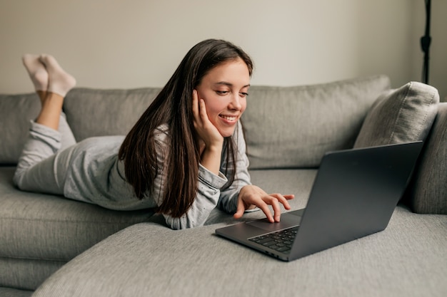 Jolie jeune femme européenne aux longs cheveux noirs portant des pyjamas travaillant à la maison avec un ordinateur portable pendant la quarantaine.