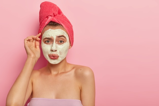 Jolie jeune femme avec une expression gênée, applique un masque crème sur le visage pour réduire les problèmes de peau, a un regard mécontent, prend un bain tous les jours, bénéficie de procédures hygiéniques. Soins de santé