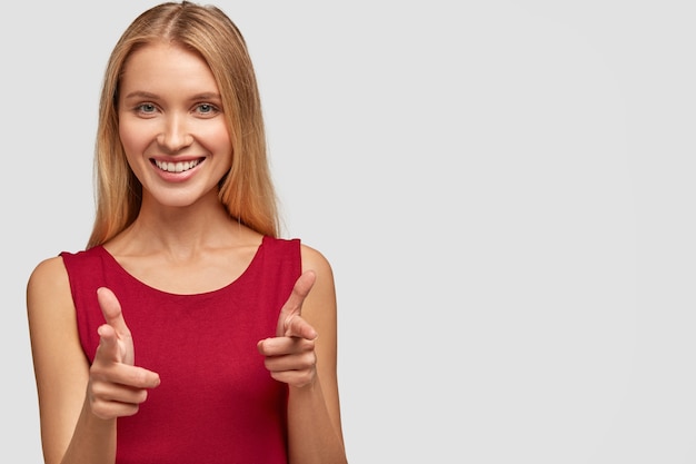 Jolie jeune femme avec une expression heureuse, vous montre du doigt ou fait des coups de feu avec les doigts, porte un gilet décontracté rouge