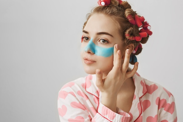 Jolie jeune femme féminine en bigoudis et vêtements de nuit appliquer un masque facial